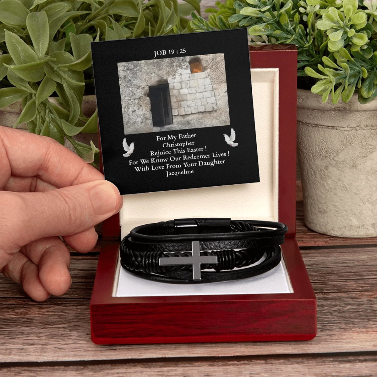 Black leather bracelet with cross charm in a red box, held by a hand with plants in the background.