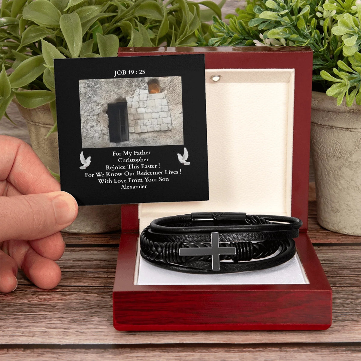 Black leather bracelet with cross charm in a red box, held by a hand with plants in the background.