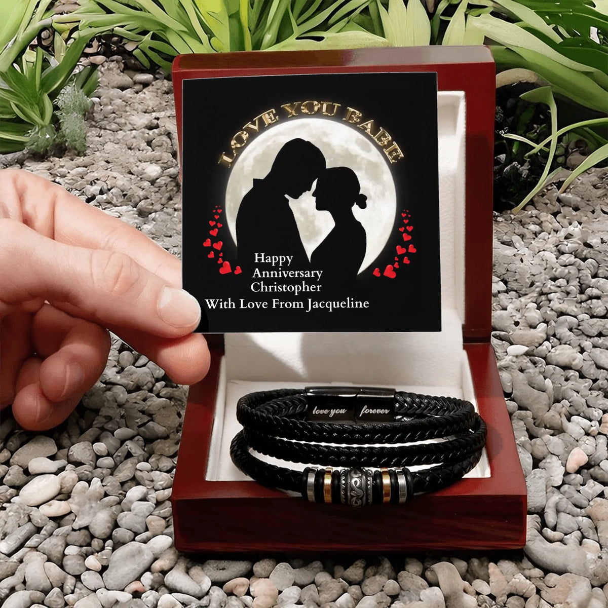 A hand holds an open wooden gift box containing a black braided bracelet with a silver-toned engraved centerpiece. The box is lined with white fabric and displays a black memorial card with a couple silhouette and the text 'I LOVE YOU BABE Happy Anniversary Christopher With Love From Jacqueline.