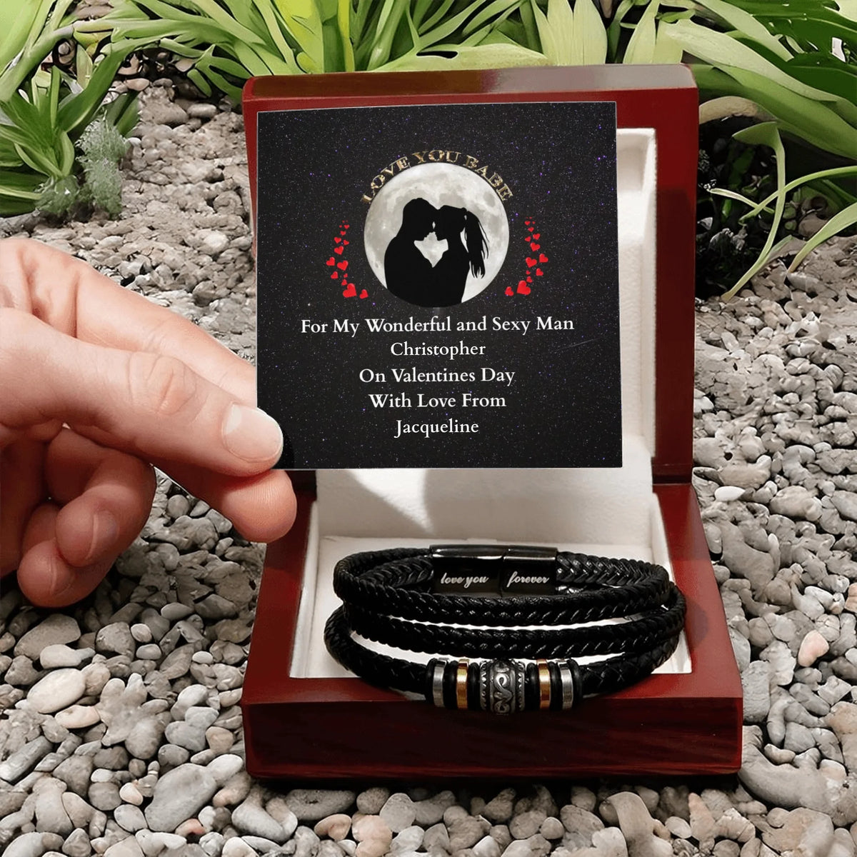Two leather bracelets in a wooden box with a personalized card on pebbles.