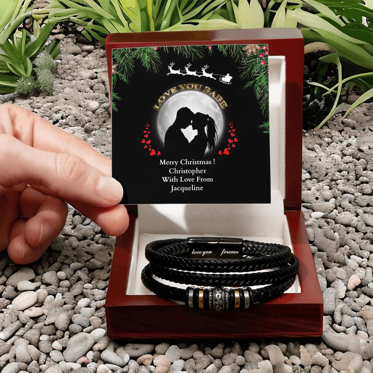 Two black leather bracelets with metal clasps in a wooden box with a Christmas-themed card on a pebbled surface.