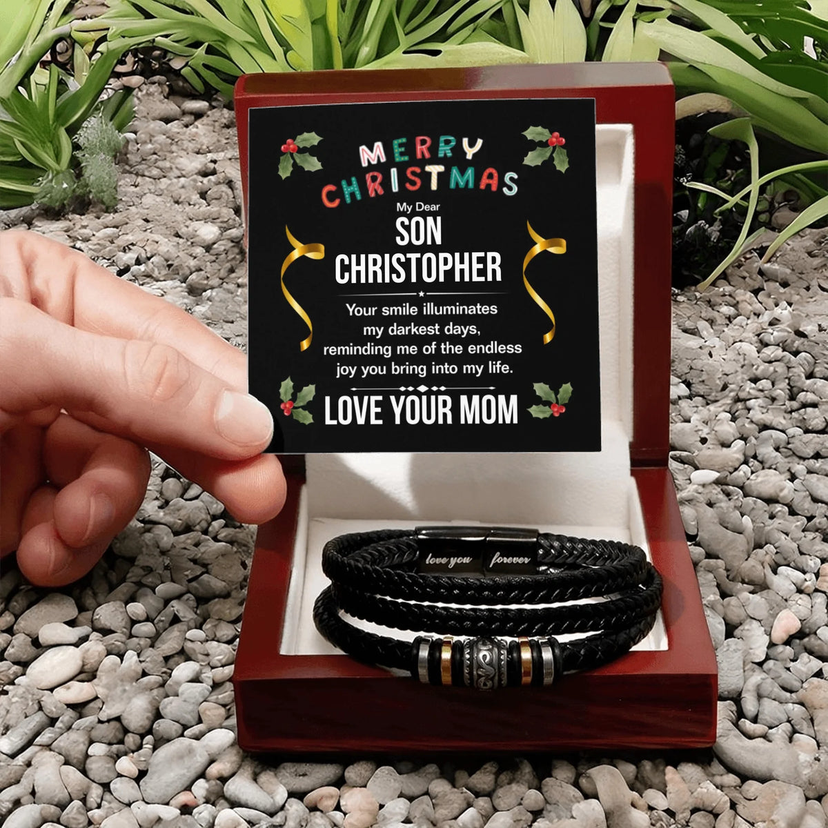 Personalized bracelet set in a decorative box with a Christmas message, held by a hand on a pebbled surface.