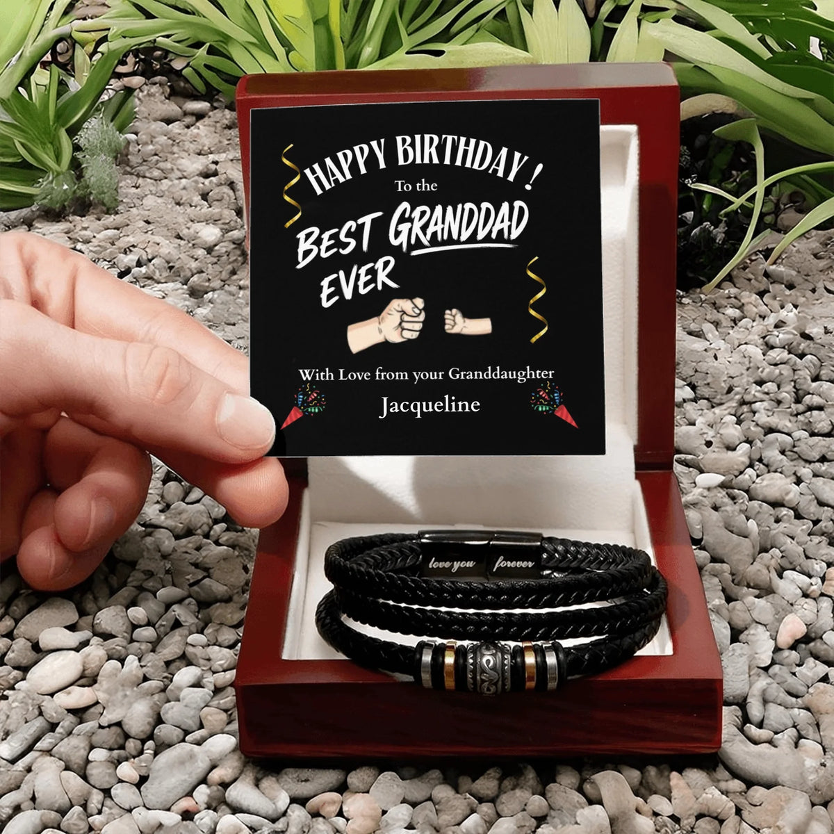 Gift box with leather bracelets and a personalized card on a pebbled surface with plants.