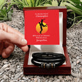 Two black leather bracelets in a wooden box with a red card on a pebbly surface.