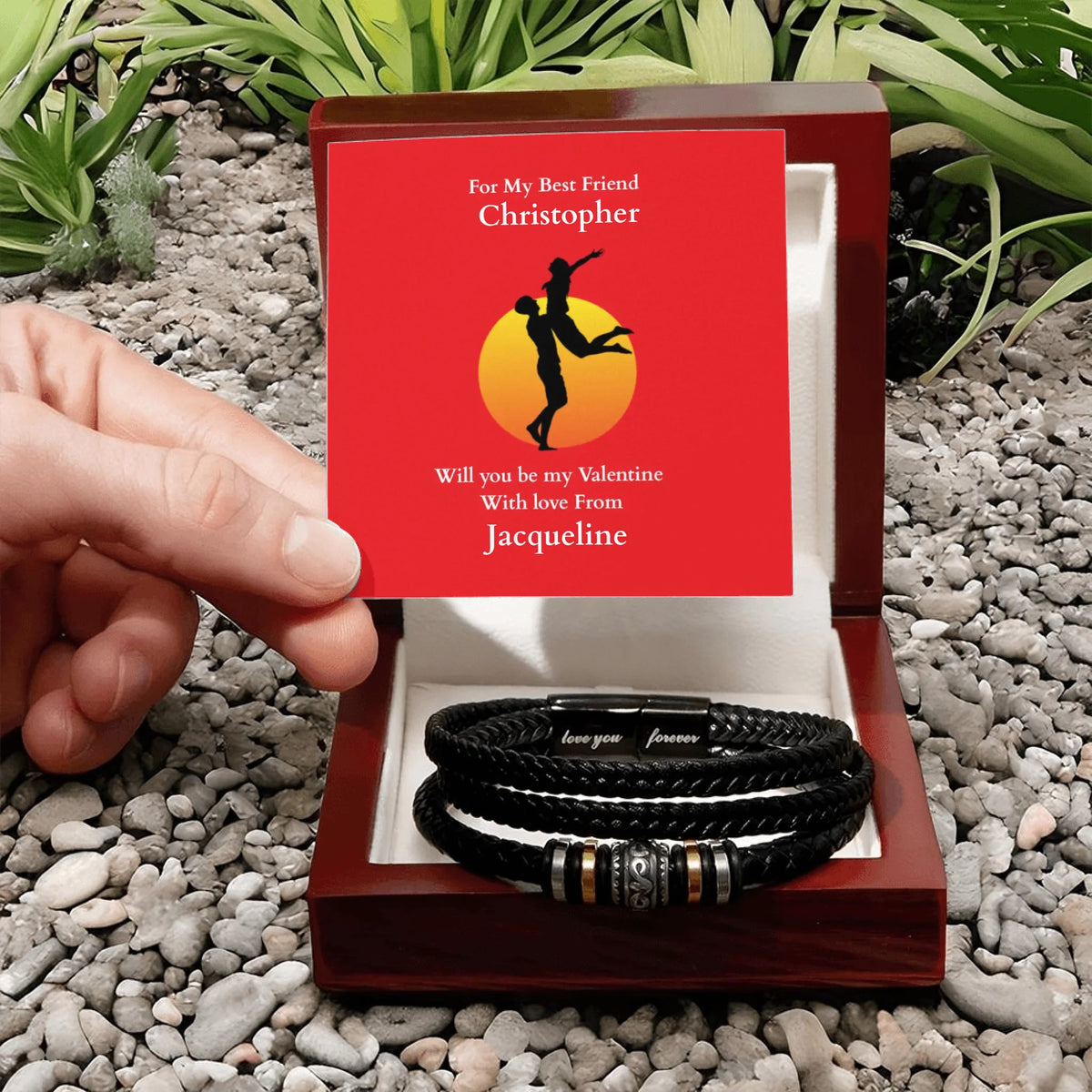 Two black leather bracelets in a wooden box with a red card on a pebbly surface.
