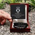 Two leather bracelets in a wooden box with a personalized message card on a pebbled surface.