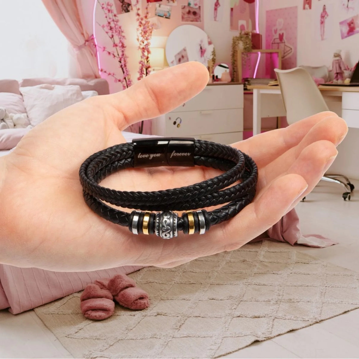 A black braided leather bracelet with silver and gold metallic accents, held in a person's hand against a pink bedroom background.