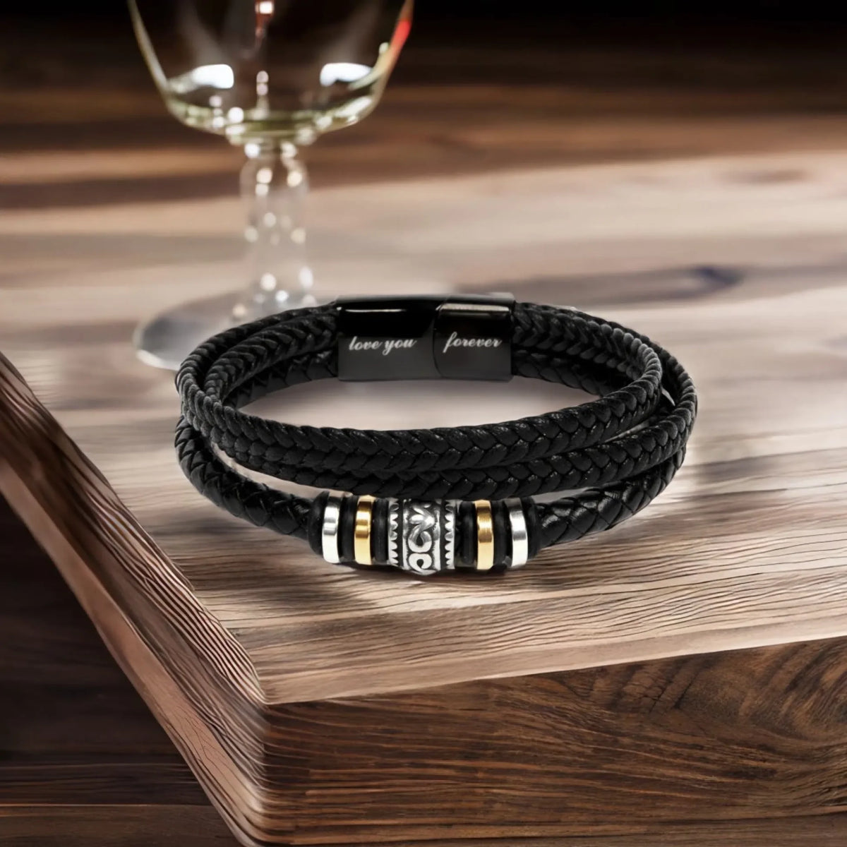 A black braided leather bracelet with a silver and gold striped magnetic clasp, resting on a wooden surface with a wine glass in the background.