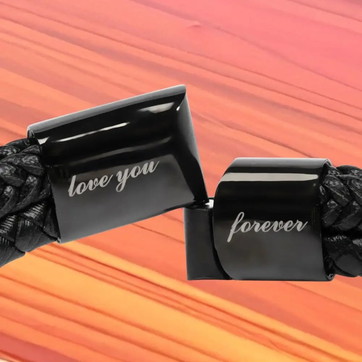 Close-up of a black braided cord with two glossy black rectangular clasps engraved with the words 'love you' and 'forever' in white script, set against a warm orange and pink striped background.