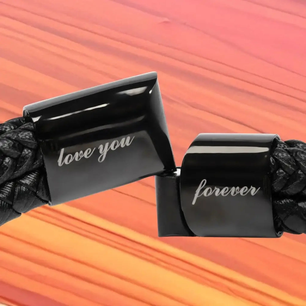 Close-up of a black braided cord with two glossy black rectangular clasps engraved with the words 'love you' and 'forever' in white script, set against a warm orange and pink striped background.