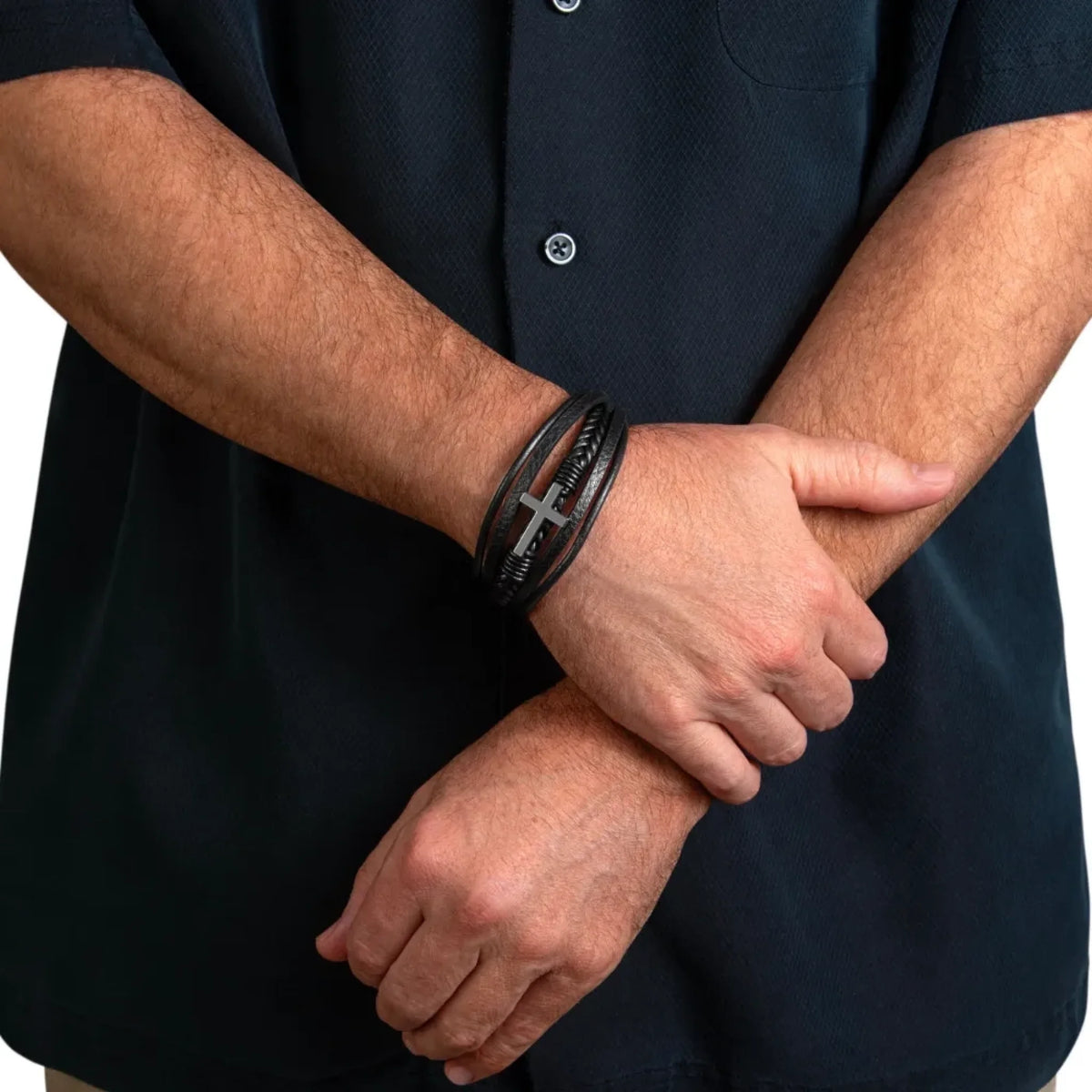 A close-up of a man's crossed arms wearing a black leather bracelet with a silver cross and engraved text on one wrist, paired with a dark polo shirt.